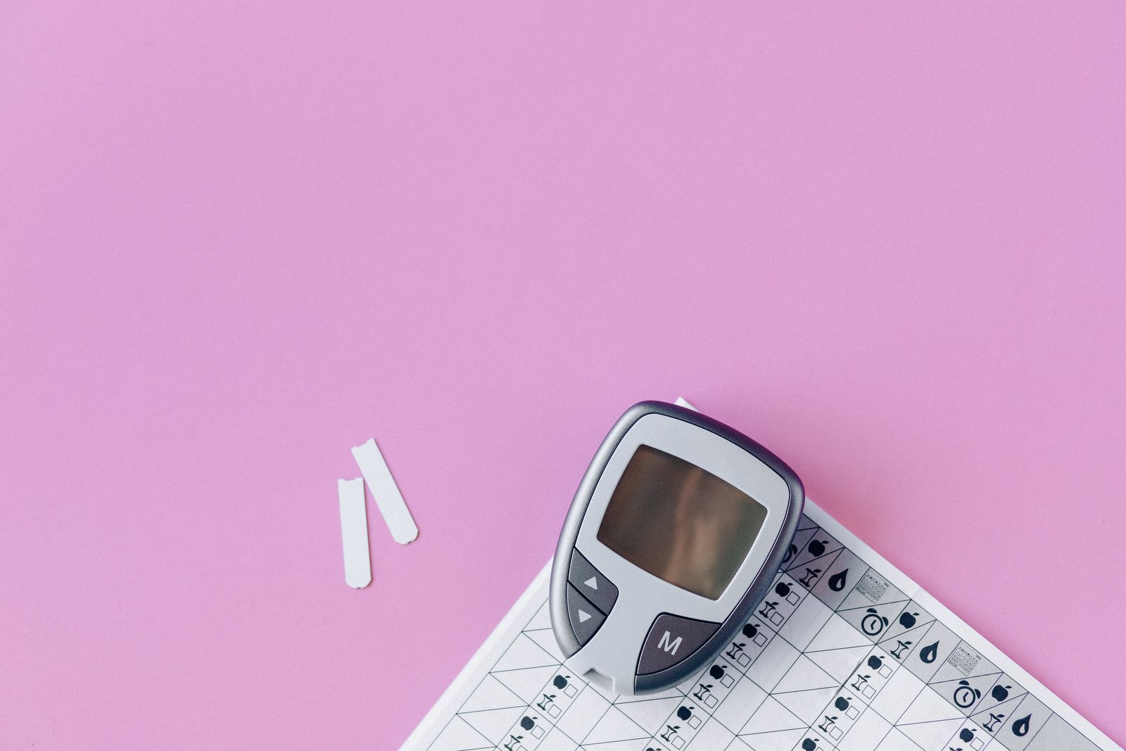 Grey blood glucose meter beside two test strips and a printed glucose log on a pink surface